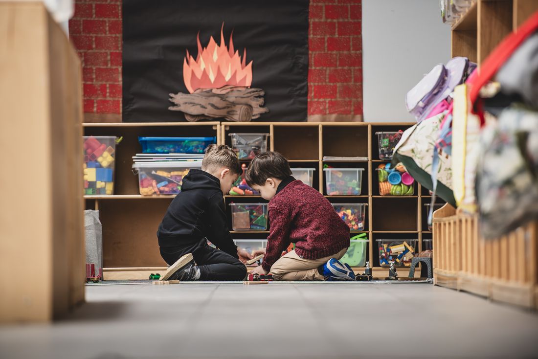 Two boys play on the floor in front of a low cubby unit with a felt campfire display above it