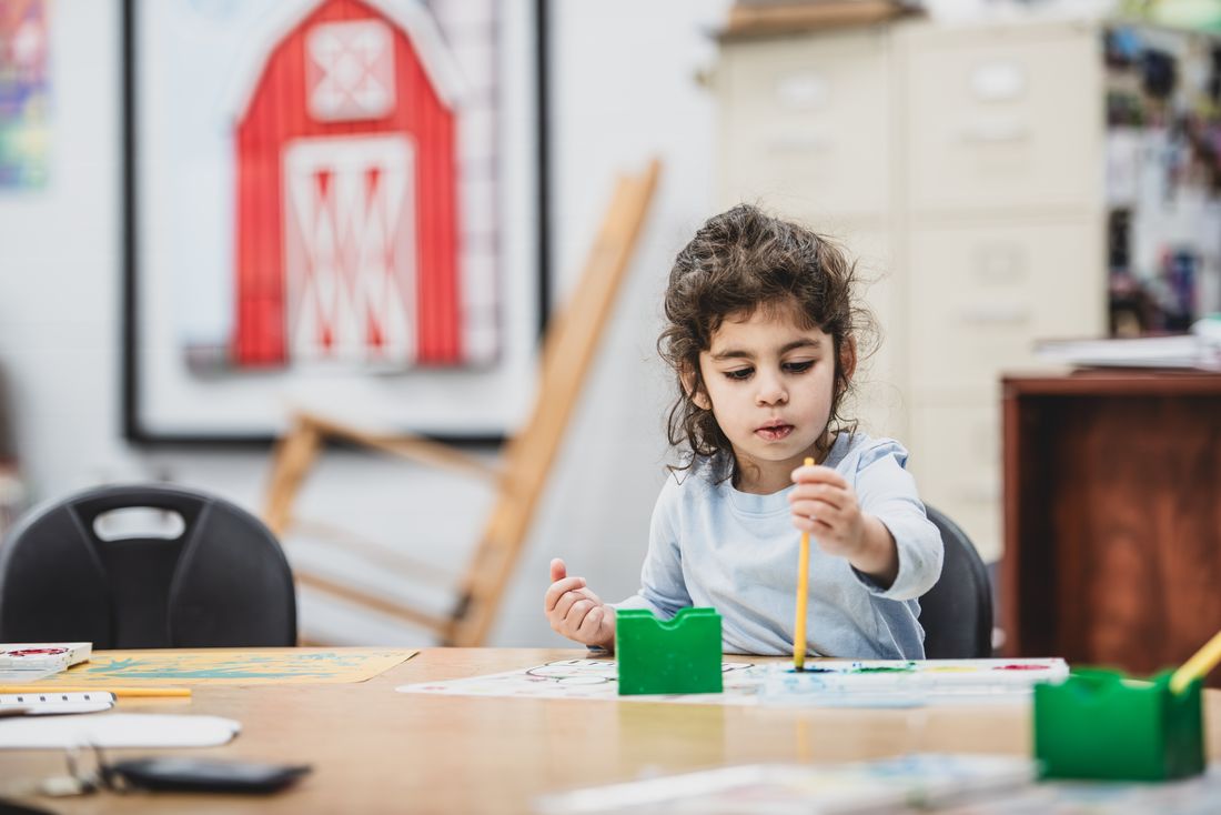 A preschooler paints at a table with green paint cups and a red barn poster behind her