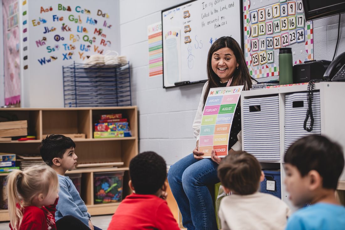 A teacher holds up a colorful chart and smiles at her class