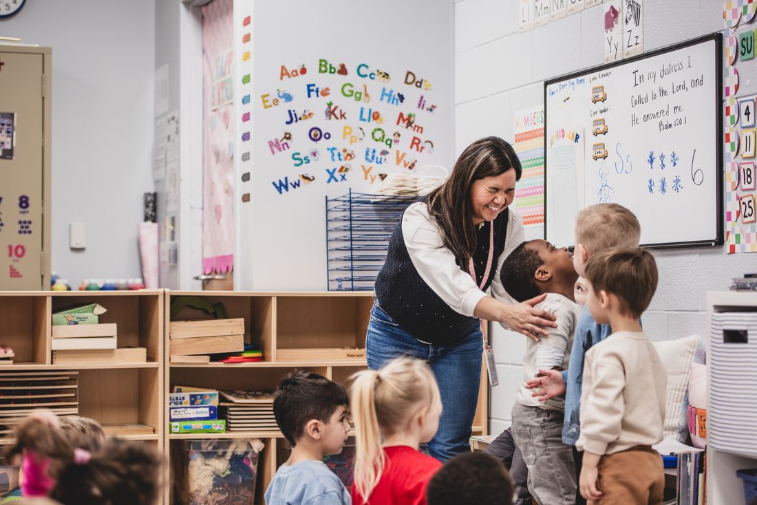 A teacher laughs and reaches to hug a boy while his classmates gather around her