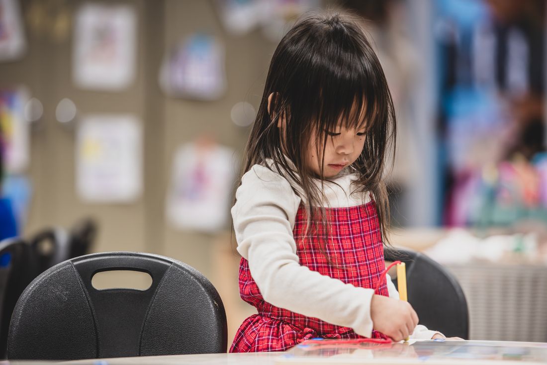 A girl in a red plaid pinafore works quietly at a table