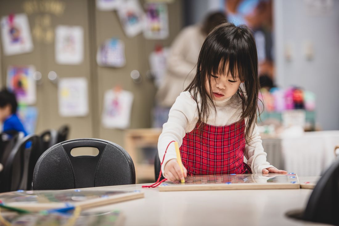 A girl in a red plaid pinafore holds a pencil and concentrates on her work