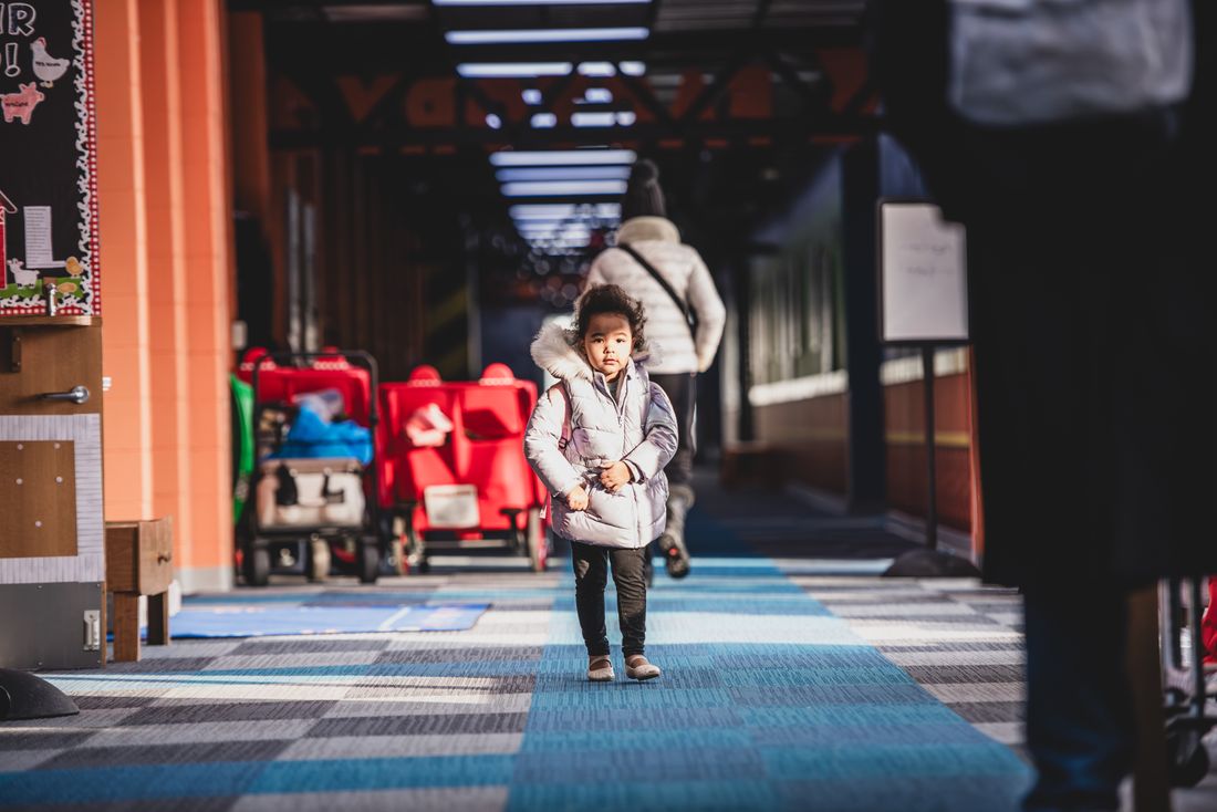 A small girl in a grey coat walks alone down the long school hallway toward the camera