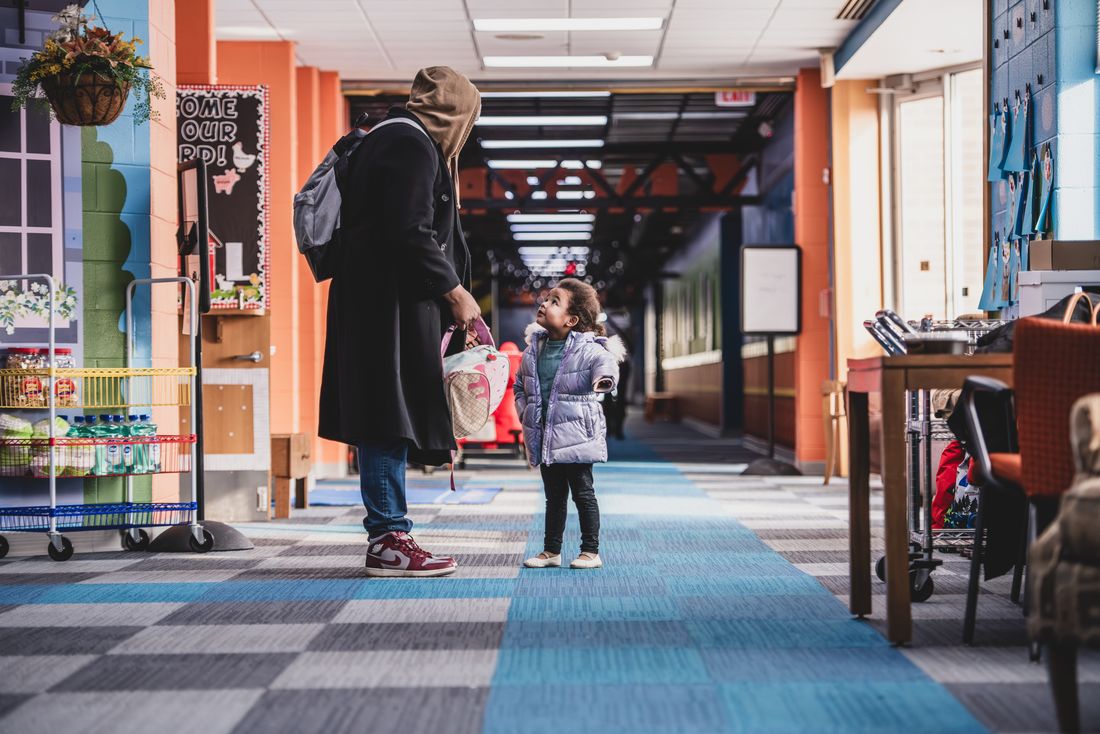 A parent and a small girl in a lavender coat look at each other in the long school hallway, a blue carpet path leading ahead