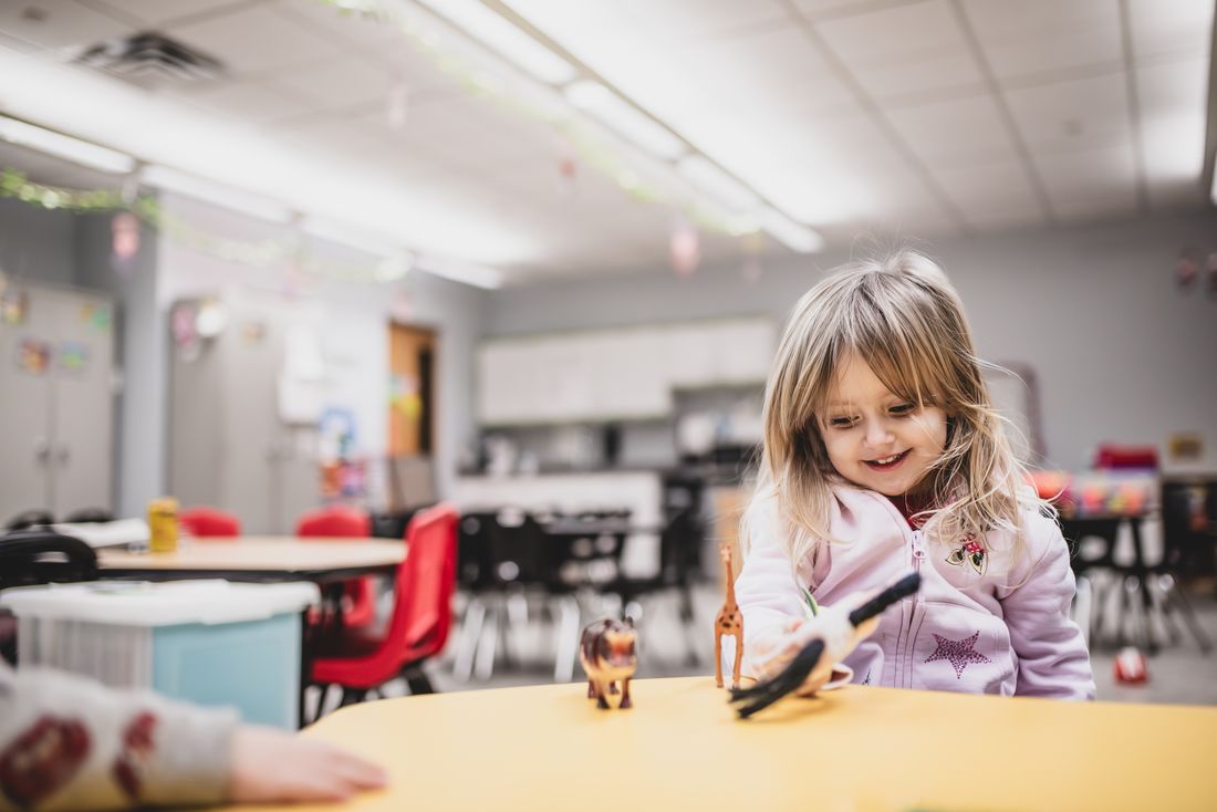 A smiling preschooler in a pink hoodie reaches for toy animals on a bright yellow table in a Train Station classroom