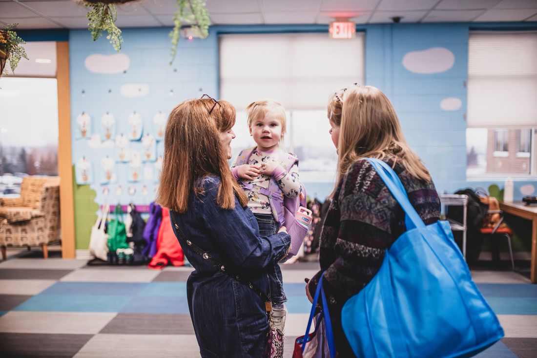 A mother holds her toddler while chatting with a teacher in a room with sky-blue cloud walls