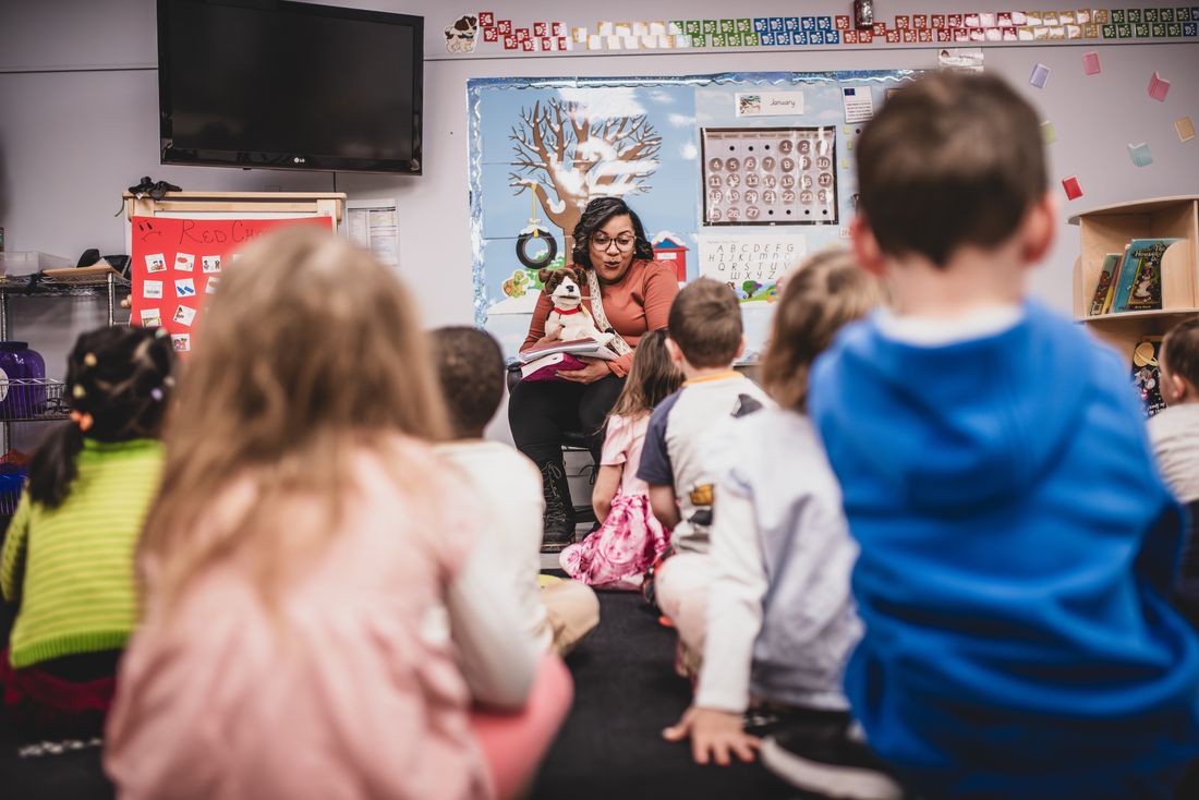 Children sit on the classroom rug facing their teacher, who reads with a puppet at the front