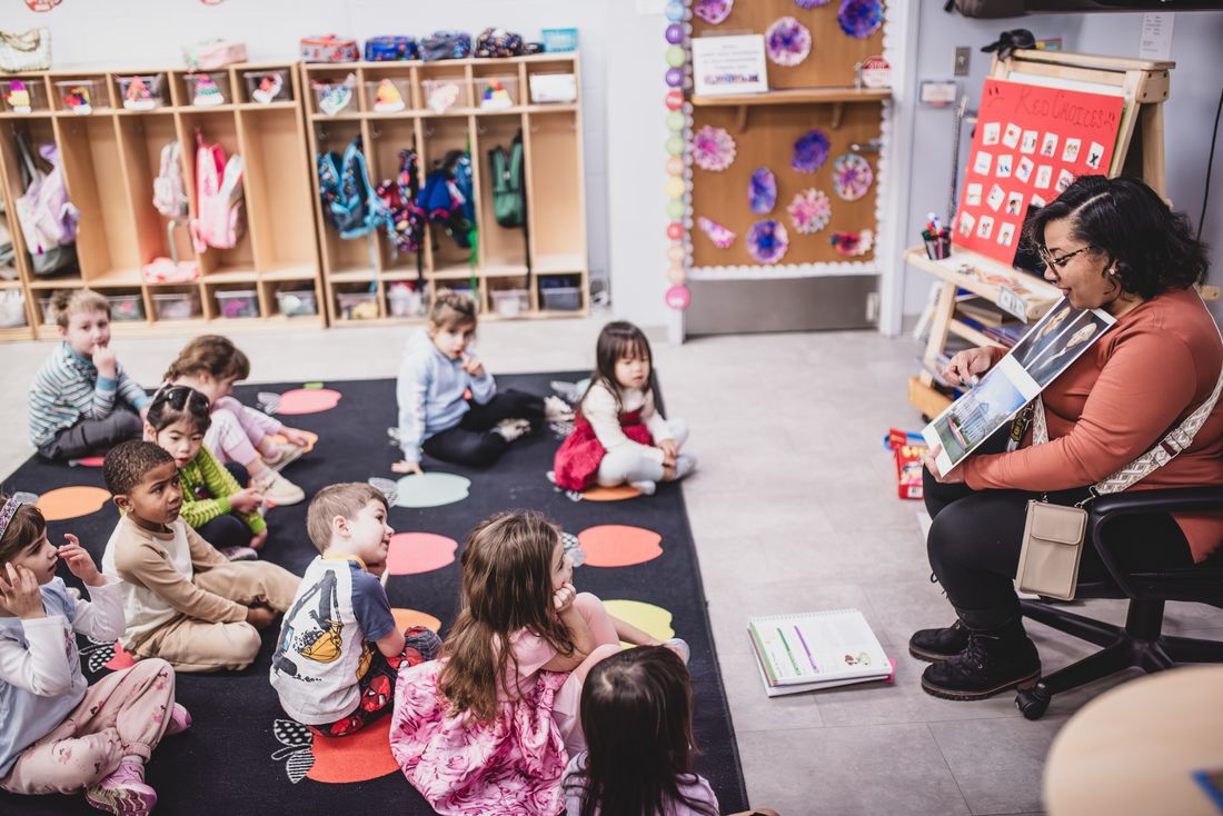 A teacher reads a picture book to children sitting on a rug with coloured dots