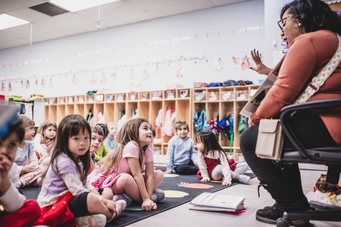 A teacher reads to a circle of preschoolers who look up from a rug with colored dots