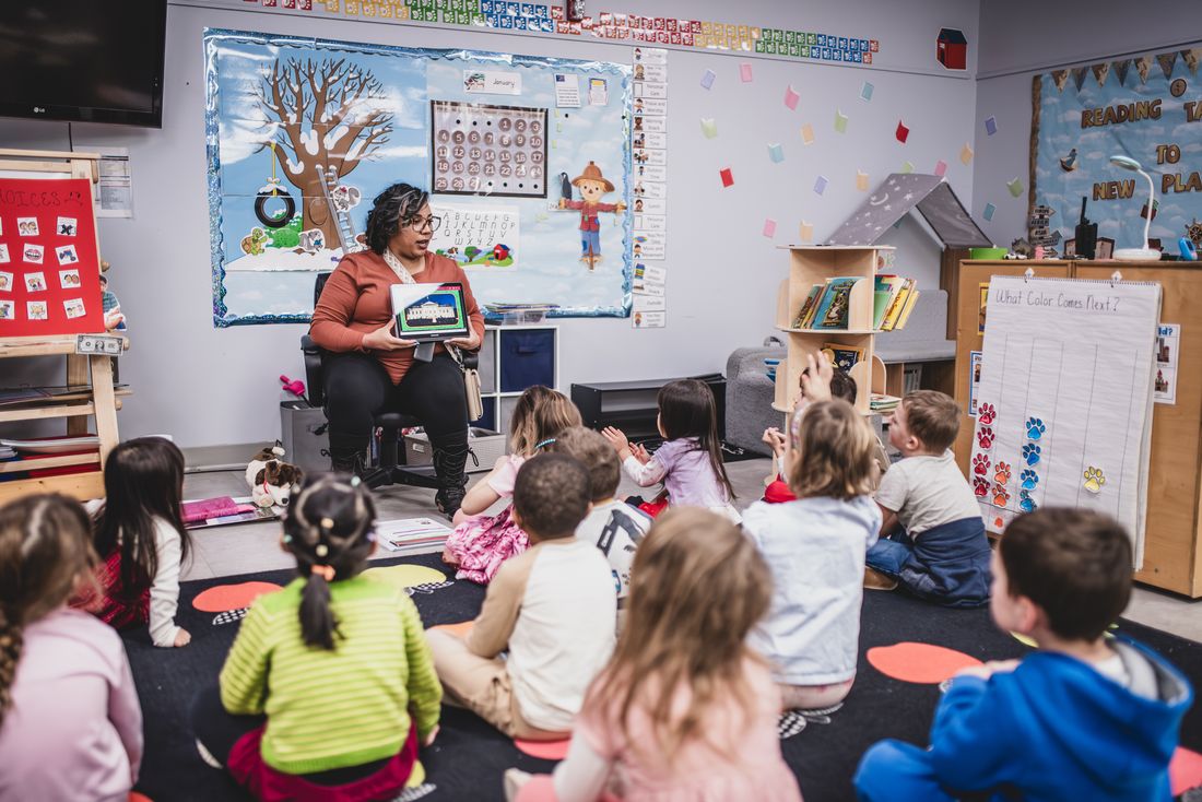 A teacher reads to her class from a chair at the front while the children sit on the rug