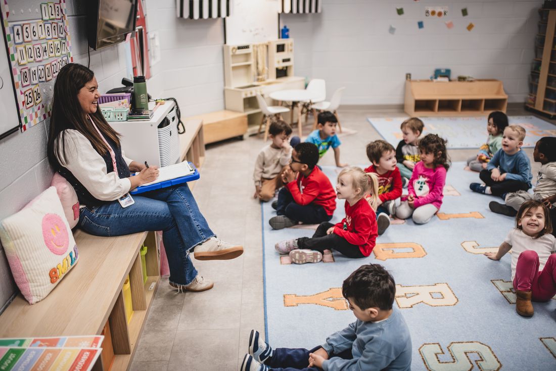 A teacher with a clipboard sits on a bench while her class sits on an alphabet rug