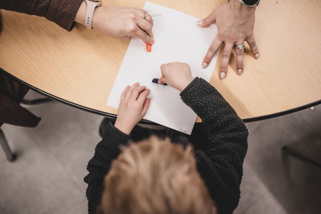 Seen from above, a teacher's hands guide a child's hand as she draws on paper