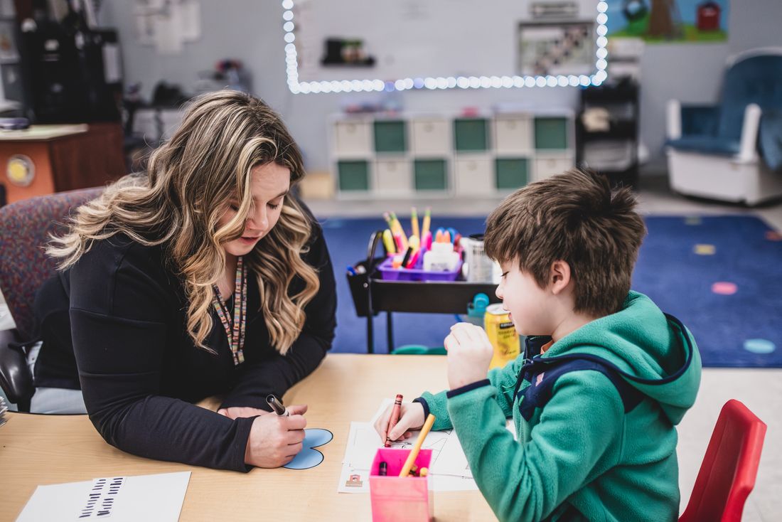 A teacher sits across a small table from a boy in a green hoodie, working with him one on one