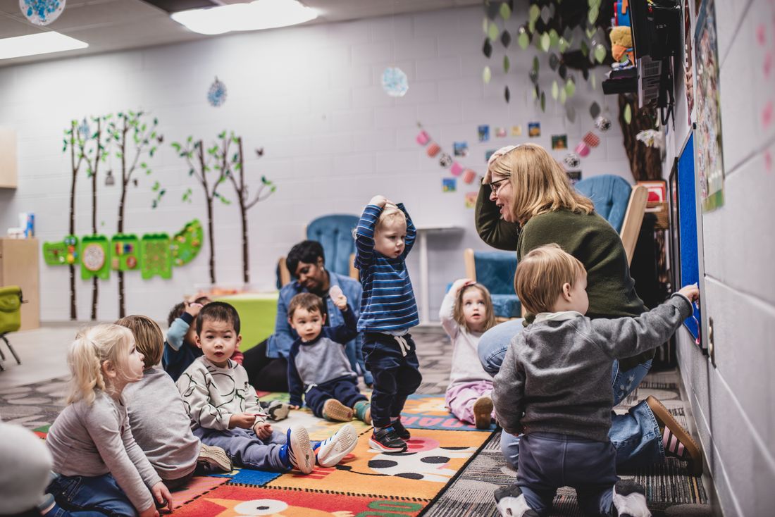 Toddlers on a colorful rug in a room with a painted tree, one standing with arms raised