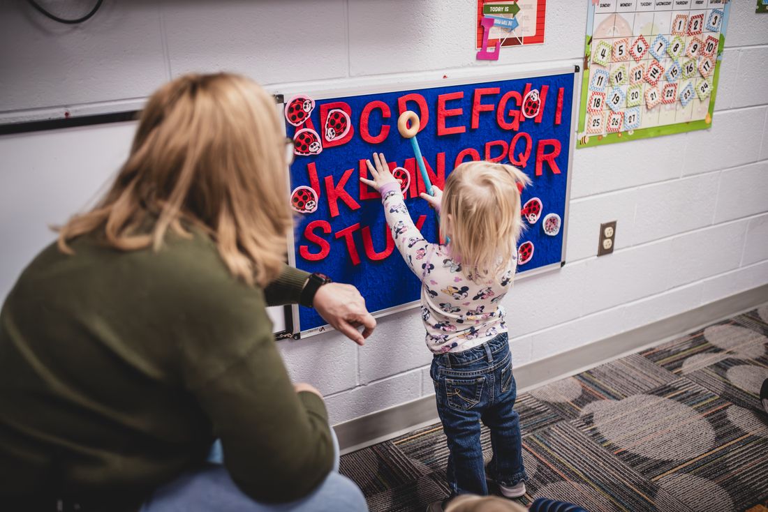 A toddler reaches up to place felt letters on a blue alphabet board while a teacher watches