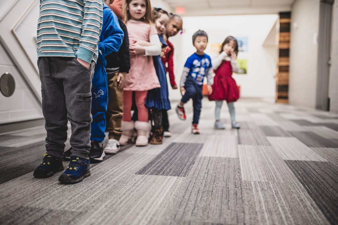 A line of preschoolers walks down the school hallway toward the camera